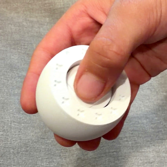 A hand pressing the centre of the Orban Clicker keychain, thumb on the stamped cap, showing its size against the fingers
